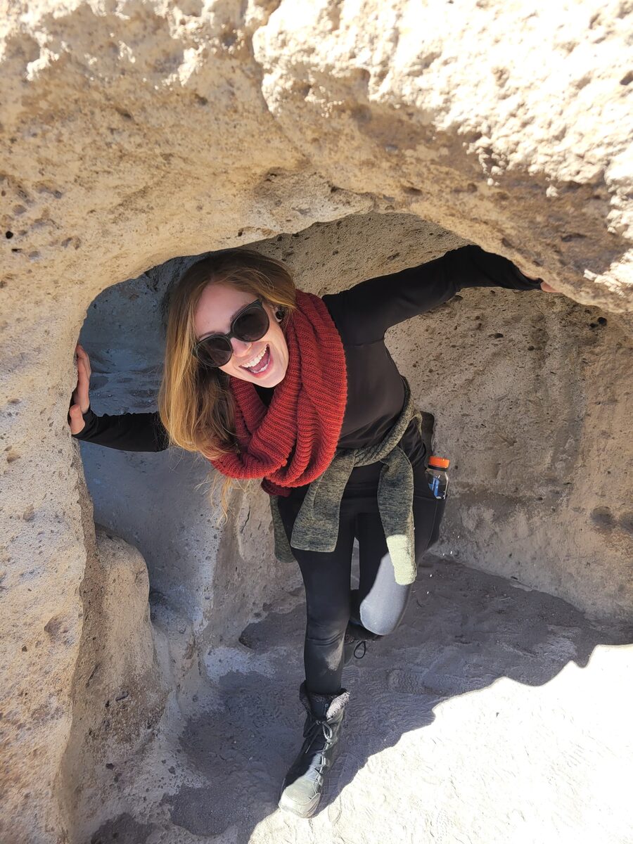 Natasha smiling in sunglasses and a red scarf inside a sandstone rock arch